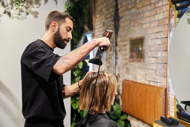 Side view of a bearded hiptser hairdresser drying a woman's hair with a blow dryer at the beauty center.